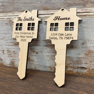 May include: Two wooden key-shaped ornaments with house designs. The first ornament says "The Smiths" and "First Christmas In Our New Home 2022". The second ornament says "Home" and "1234 Oak Lane Dallas, TX 75874".