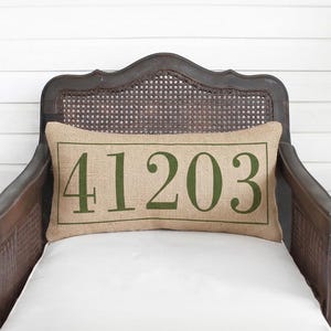 May include: A rectangular burlap pillow with the number "41203" printed in green. The pillow is placed on a vintage wooden chair with a woven back and white cushion. The background is a white wooden wall.