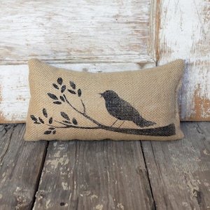 May include: A rectangular burlap pillow with a black silhouette of a bird perched on a branch. The pillow is tan and has a rustic, textured appearance. The bird and branch design is centered on the pillow.