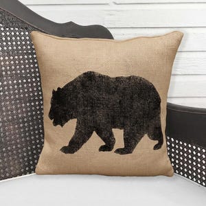 May include: Square beige burlap pillow with a black bear silhouette. The pillow is placed on a dark wooden chair with a white wooden background. The bear design is a rustic, textured print.