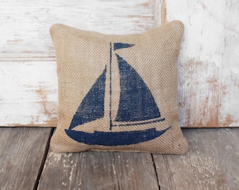 May include: Square burlap pillow with a navy blue sailboat design. The pillow is beige and has a textured surface. The sailboat graphic features a mast, sail, and hull. The pillow is placed on a wooden surface.