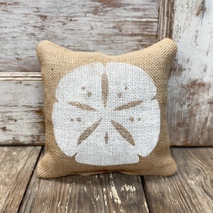 Könnte beinhalten: Ein quadratisches Dekokissen aus beigefarbenem Jute-Stoff. In der Mitte des Kissens befindet sich ein großer, weißer Siebdruck einer Sanddollar-Grafik, die das charakteristische fünfzackige Sternmuster und kleine kreisförmige Details zeigt.