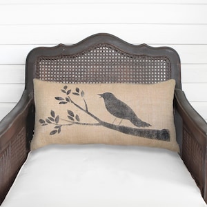 May include: A rectangular lumbar pillow made of tan burlap fabric featuring a black screen-printed graphic of a bird perched on a leafy branch. The pillow is positioned horizontally against the backrest of a dark brown wooden chair.
