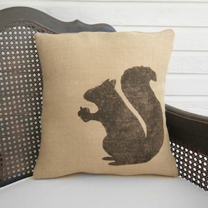 May include: A square throw pillow made of tan, textured burlap fabric. The center of the pillow features a dark brown, solid silhouette graphic of a squirrel sitting upright and holding a small acorn in its front paws, facing to the left.
