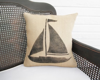 May include: A square throw pillow made of a light tan, textured fabric featuring a dark charcoal gray screen-printed graphic of a sailboat. The design shows the hull, mast, and two triangular sails of the boat centered on the front of the pillow.
