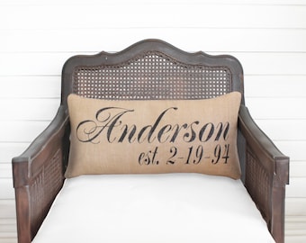 May include: A rectangular tan burlap pillow featuring the black printed text Anderson and est. 2-19-94 in a decorative script and serif font. The pillow is positioned horizontally against the backrest of a dark brown wooden chair with cane detailing.