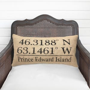 May include: A rectangular burlap pillow with black text reading "46.3188° N 63.1461° W Prince Edward Island." The pillow is on a vintage wooden chair with a woven back and white cushion.