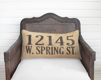 May include: A rectangular tan burlap lumbar pillow featuring black printed text that reads 12145 W. SPRING ST. The pillow is positioned on a dark brown wooden chair with a woven cane backrest and a plain white seat cushion.