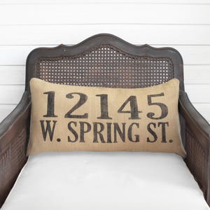 May include: A rectangular burlap throw pillow featuring the black printed text 12145 W. SPRING ST. centered on the front. The pillow is positioned against the backrest of a dark brown wooden chair with a woven cane back panel and white seat.