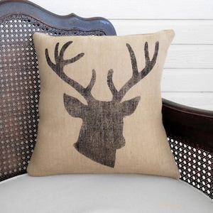 May include: A square burlap pillow featuring a dark brown silhouette of a deer head with antlers. The pillow is resting on a white cushion, with a dark brown wooden chair in the background. The pillow has a rustic, farmhouse style.