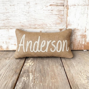 May include: A rectangular burlap pillow with the name "Anderson" printed in white script. The pillow is centered on a weathered wooden surface, with a distressed white wooden background.