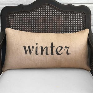 May include: A rectangular lumbar pillow made of tan, textured burlap fabric. The word winter is printed in the center in a black, serif-style font. The pillow has a simple, clean design with visible woven fibers throughout the material.