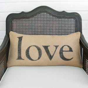 May include: A rectangular tan burlap pillow featuring the word love printed in large black lowercase letters. The pillow is positioned horizontally against the dark wood and cane backrest of a chair with a white seat cushion.