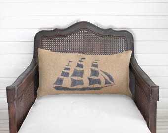 May include: A rectangular lumbar pillow made of tan burlap fabric featuring a dark blue screen-printed graphic of a sailing ship with multiple masts and sails centered on the front surface of the pillow cover.