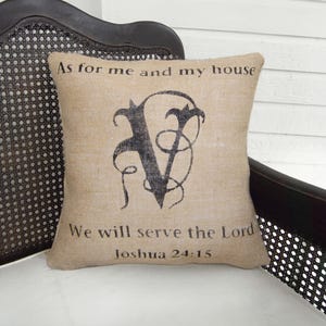 May include: Square burlap pillow with black text and a decorative letter "V". The text reads "As for me and my house, We will serve the Lord, Joshua 24:15". The pillow is on a white surface, with a dark brown chair in the background.