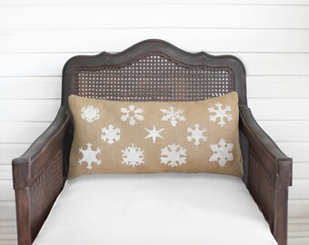 May include: A rectangular lumbar pillow made of tan burlap fabric featuring a repeating pattern of white printed snowflakes in various shapes and sizes across the front surface, centered on a dark brown wooden chair with a cane backrest.