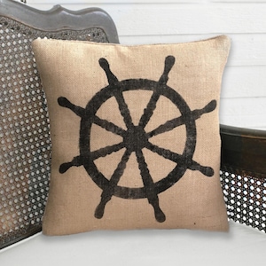 May include: Square burlap pillow with a black ship's wheel design. The pillow is tan with a dark, hand-painted nautical wheel. The pillow is placed on a white surface, with a vintage chair in the background.