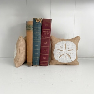 May include: Three vintage hardcover books standing upright between two small square burlap pillows. The books are tan, blue, and maroon with visible text on their spines. Each burlap pillow features a white printed sand dollar design.