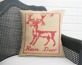 May include: A square burlap throw pillow featuring a red screen-printed graphic of a deer with large antlers. The design is enclosed in a rectangular border with the number 5 in the top right corner and the text Rein Deer printed at the bottom.