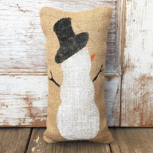 May include: A rectangular burlap pillow featuring a snowman design. The snowman has a black top hat, a white body, an orange nose, and stick arms. The pillow is a festive winter decoration.