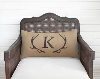May include: A rectangular tan burlap lumbar pillow featuring a dark brown printed design of a capital letter K centered between two symmetrical deer antlers. The pillow is positioned on a dark brown wooden chair with a woven cane backrest.