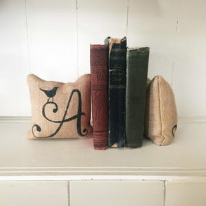 May include: Two tan burlap bookends with a black printed letter A and a small bird perched on top, positioned on either side of three vintage hardcover books with red, black, and dark green spines standing upright in a row.