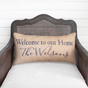 May include: A rectangular burlap pillow with the text "Welcome to our Home" and "The Wilsons" in dark blue script. The pillow rests on a white cushion within a dark brown wooden chair with a woven back.