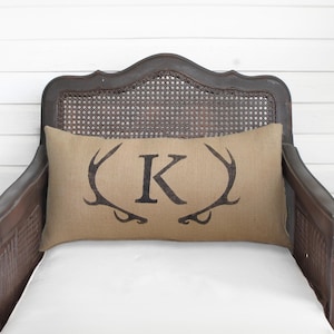 May include: A rectangular tan burlap pillow featuring a dark brown printed design of a large letter K centered between two symmetrical deer antlers. The pillow is positioned on a dark brown wooden chair with a woven cane backrest.