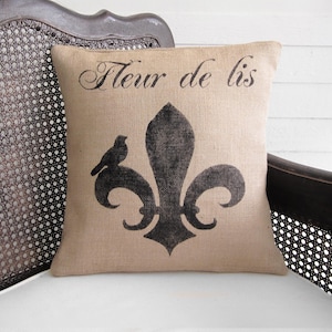 May include: A square burlap pillow with a black fleur-de-lis design and the words "Fleur de lis" printed across the top. A small bird is perched on the design. The pillow is resting on a white surface.