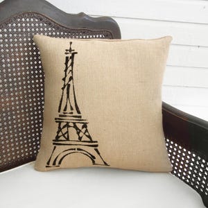 May include: A square beige burlap pillow with a black Eiffel Tower design. The pillow is placed on a dark wooden chair with a woven back. The pillow is a decorative item.