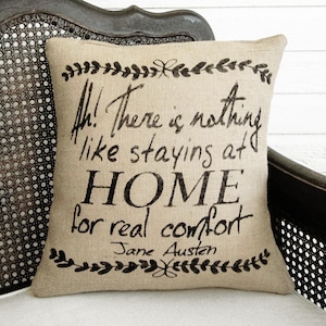 May include: A square beige burlap throw pillow featuring black printed text that reads, Ah! There is nothing like staying at HOME for real comfort, followed by the name Jane Austen, framed by decorative black leaf borders at the top and bottom.