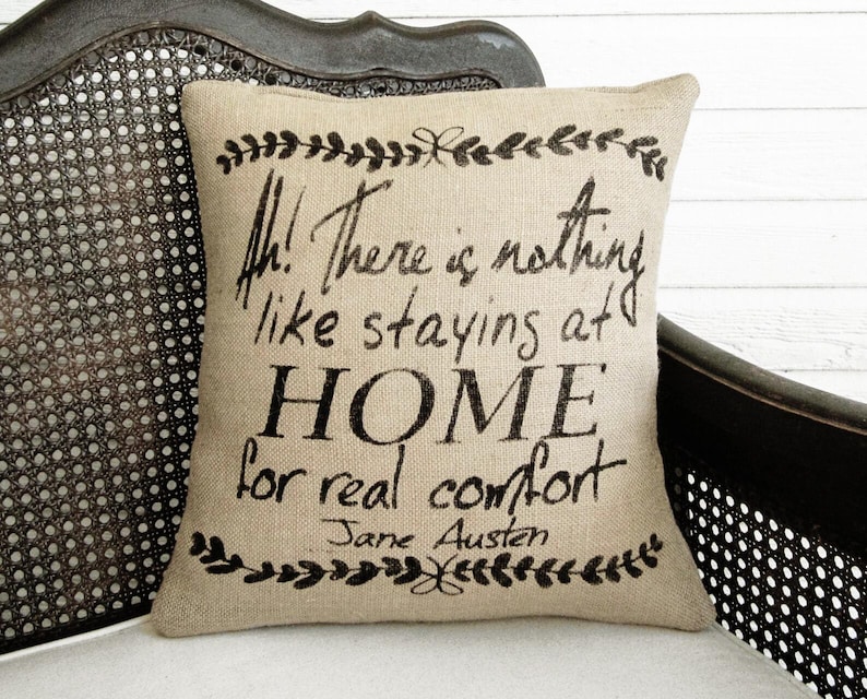 May include: A square beige burlap throw pillow featuring black printed text that reads, Ah! There is nothing like staying at HOME for real comfort, followed by the name Jane Austen, framed by decorative black leaf borders at the top and bottom.