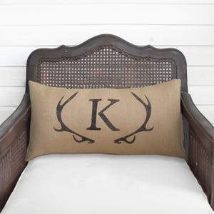May include: A rectangular burlap pillow with a dark brown antler design and the letter "K" in the center. The pillow is placed on a white cushion inside a dark wooden chair with a woven back.