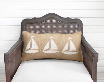May include: A rectangular lumbar pillow made of tan burlap fabric featuring a repeating white screen-printed graphic of three sailboats. The pillow is positioned horizontally, displaying a textured weave and a simple nautical design.