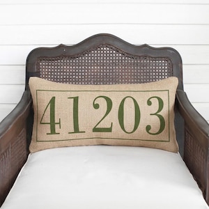 May include: A rectangular burlap throw pillow featuring the green printed numbers 41203 centered within a thin green rectangular border. The pillow has a textured, woven tan fabric surface and is positioned horizontally on a dark wooden chair.