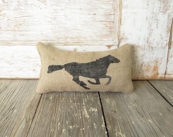 Könnte beinhalten: Ein rechteckiges Kissen aus Jute mit einer schwarzen Silhouette eines galoppierenden Pferdes auf der Vorderseite. Das Kissen hat eine grobe, gewebte Textur und ist gefüllt, um eine weiche, leicht abgerundete Form mit spitzen Ecken zu erzeugen.