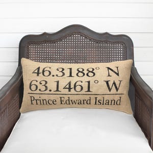 May include: A rectangular burlap throw pillow featuring black printed text that reads 46.3188° N 63.1461° W and Prince Edward Island. The pillow has a coarse, tan-colored woven texture and is positioned horizontally against a dark wooden chair back.