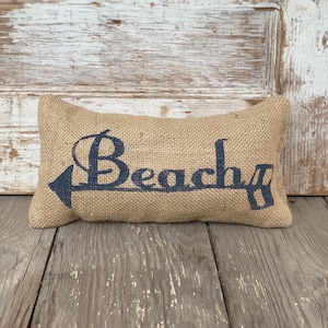 May include: A rectangular burlap pillow featuring the word Beach printed in dark blue ink. The text is stylized with an arrow pointing to the left, integrated into the first letter. The fabric has a coarse, woven texture and a natural tan color.
