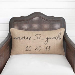 May include: A rectangular burlap pillow with the names "annie" and "jacob" in black script, joined by a heart symbol. Below the names is the date "10-20-18". The pillow rests on a white cushion within a dark wooden chair.