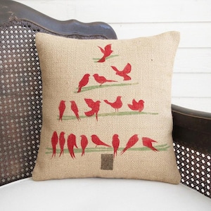 A beige pillow with a Christmas tree design made of red birds on a wooden chair.