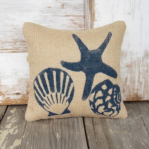 Könnte beinhalten: Ein quadratisches Kissen aus Jute mit marineblauen Muschel- und Seestern-Designs. Das Kissen hat eine natürliche beige Farbe und liegt auf einer Holzoberfläche. Das Design des Kissens erinnert an ein Küsten- oder maritimes Thema.