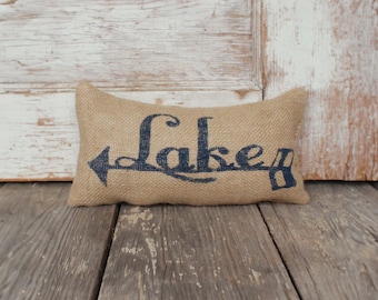 Könnte beinhalten: Ein rechteckiges Zierkissen aus Jute mit dem Wort Lake, das in dunkelblauer Tinte aufgedruckt ist. Der Schriftzug ist mit einem nach links zeigenden Pfeil gestaltet, der in das Design integriert ist. Das Kissen hat eine grobe Webstruktur und Farbe.