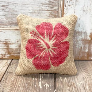 May include: A square-shaped decorative pillow made of tan, coarse-textured burlap fabric. The center of the pillow features a large, bold, red hibiscus flower print with visible stamen details, centered on the front face of the cushion.