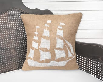 May include: A square throw pillow made of tan burlap fabric featuring a white screen-printed graphic of a sailing ship with multiple masts and sails. The pillow has a simple, clean design with a visible woven texture throughout the material.