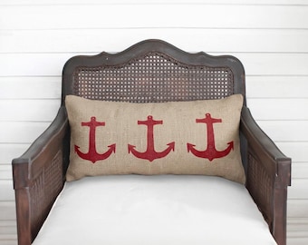 May include: A rectangular lumbar pillow made of tan burlap fabric featuring three evenly spaced, bold red anchor graphics printed across the front surface. The pillow is positioned horizontally against the backrest of a dark brown wooden chair.