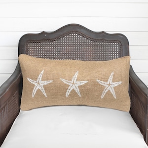 May include: A rectangular lumbar pillow made of tan, textured burlap fabric. The front of the pillow features three white, hand-painted starfish designs arranged horizontally across the center of the fabric surface.