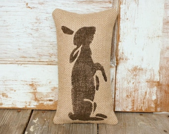 Könnte beinhalten: Ein rechteckiges Kissen aus Jute mit einer braunen Silhouette eines Hasen. Das Kissen hat eine natürliche beige Farbe und befindet sich vor einem rustikalen Holzuntergrund. Der Hase wird stehend dargestellt.