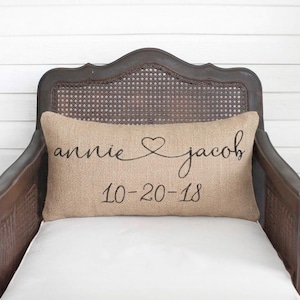 May include: A rectangular burlap throw pillow featuring the black printed text "annie" and "jacob" connected by a heart symbol, with the date "10-20-18" printed in black below the names, centered on a dark wooden chair with a cane back.