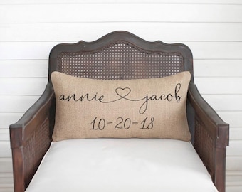 May include: A rectangular burlap throw pillow featuring the black printed text "annie" and "jacob" connected by a heart symbol, with the date "10-20-18" printed in black below the names, centered on a dark wooden chair with a cane back.