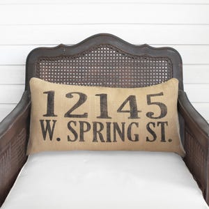 May include: A rectangular burlap pillow with the address "12145 W. SPRING ST." printed in black. The pillow rests on a white cushion in a dark wooden chair with a woven back.